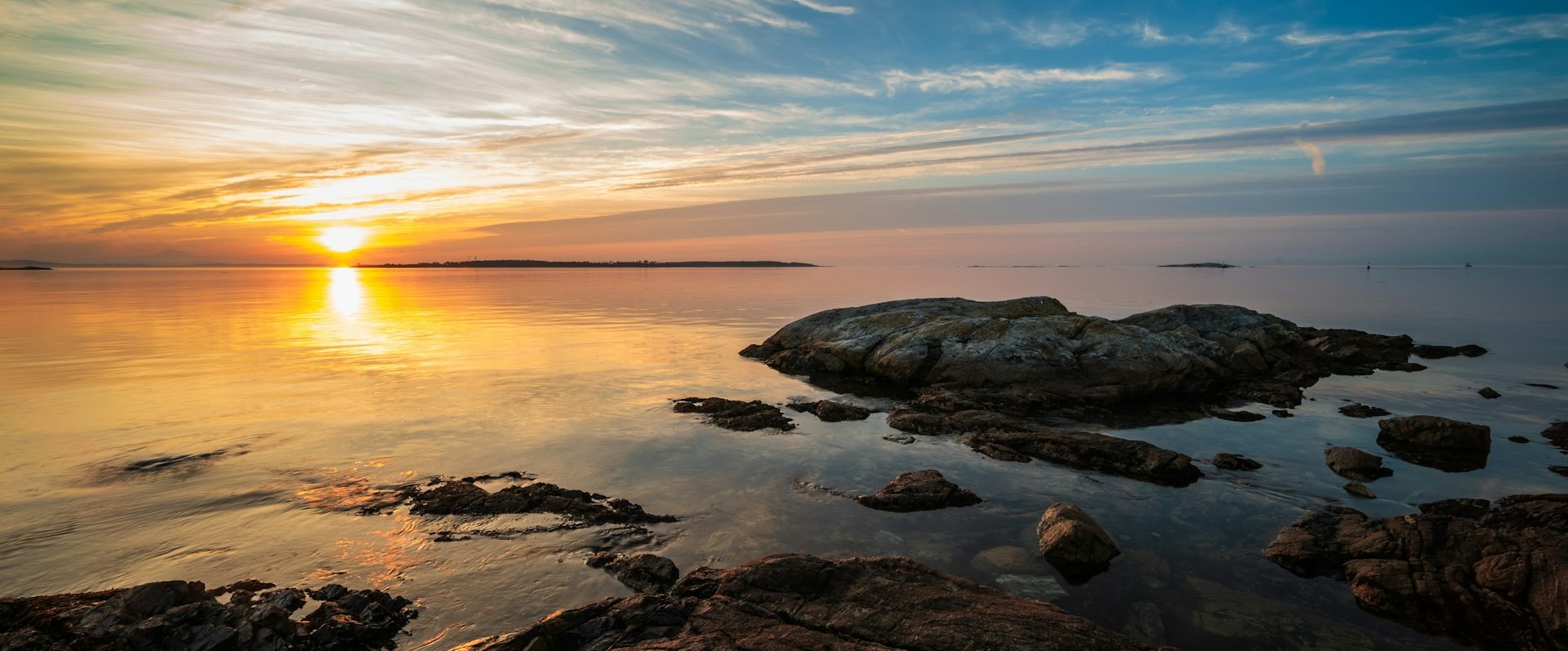 Solnedgång över stilla vatten med klippor i förgrunden.